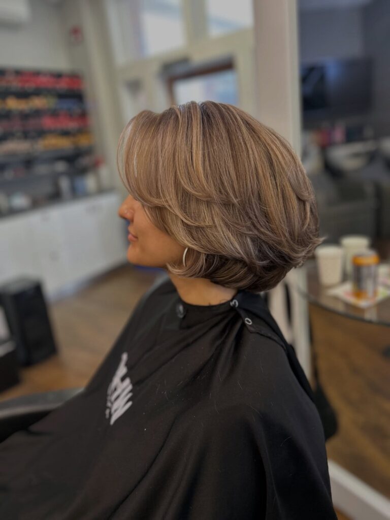 A woman in a salon chair showing a side view of her voluminous ash blonde layered bob with feathered swooping bangs.