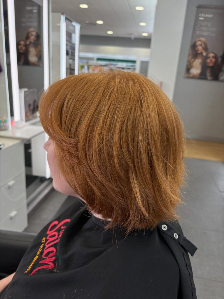 A woman in a salon chair showing a side profile of her vibrant copper-colored choppy bob with sweeping side bangs and textured layers.