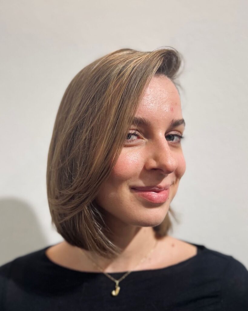 A smiling woman looking forward with a side-parted light brown and blonde choppy bob featuring a voluminous sweeping side style.