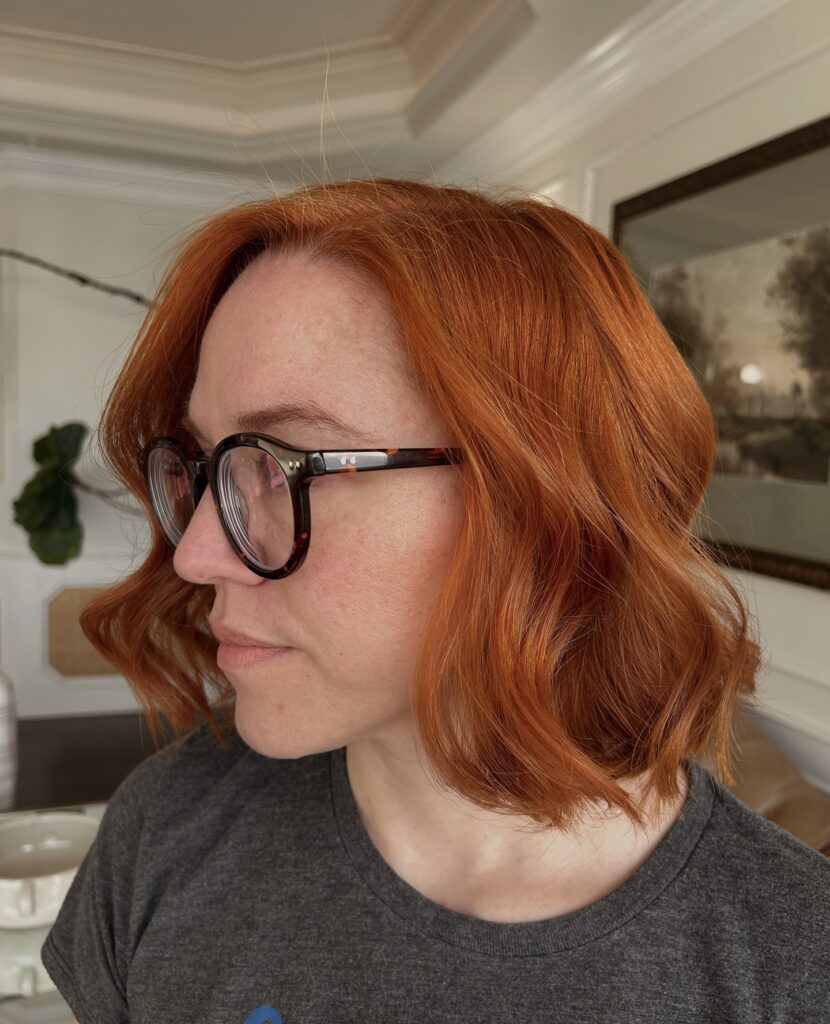 A woman with glasses viewed in side profile wearing a vibrant ginger red wavy long bob with choppy layered ends.