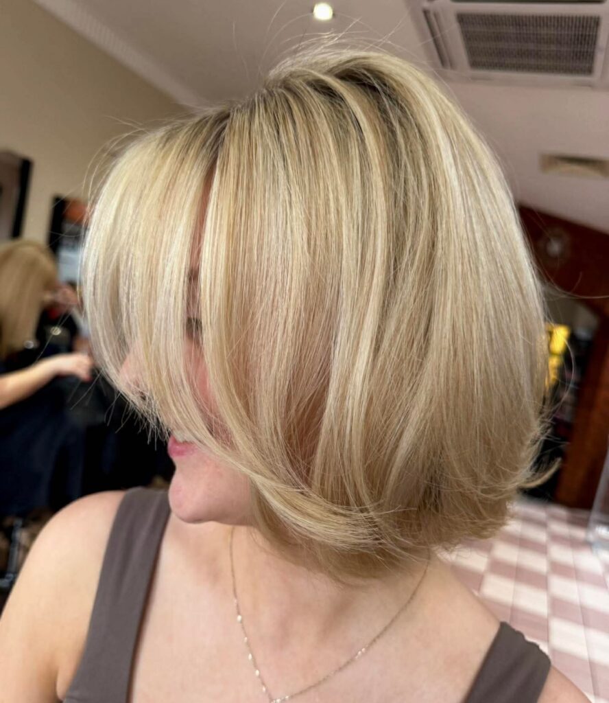 A smiling woman viewed from the side sporting a smooth creamy blonde jaw length bob with long wispy bangs framing her face.