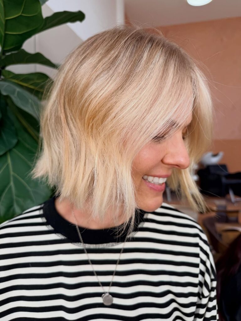 A smiling woman in side profile showing off a bright buttery blonde choppy bob with textured curtain bangs.