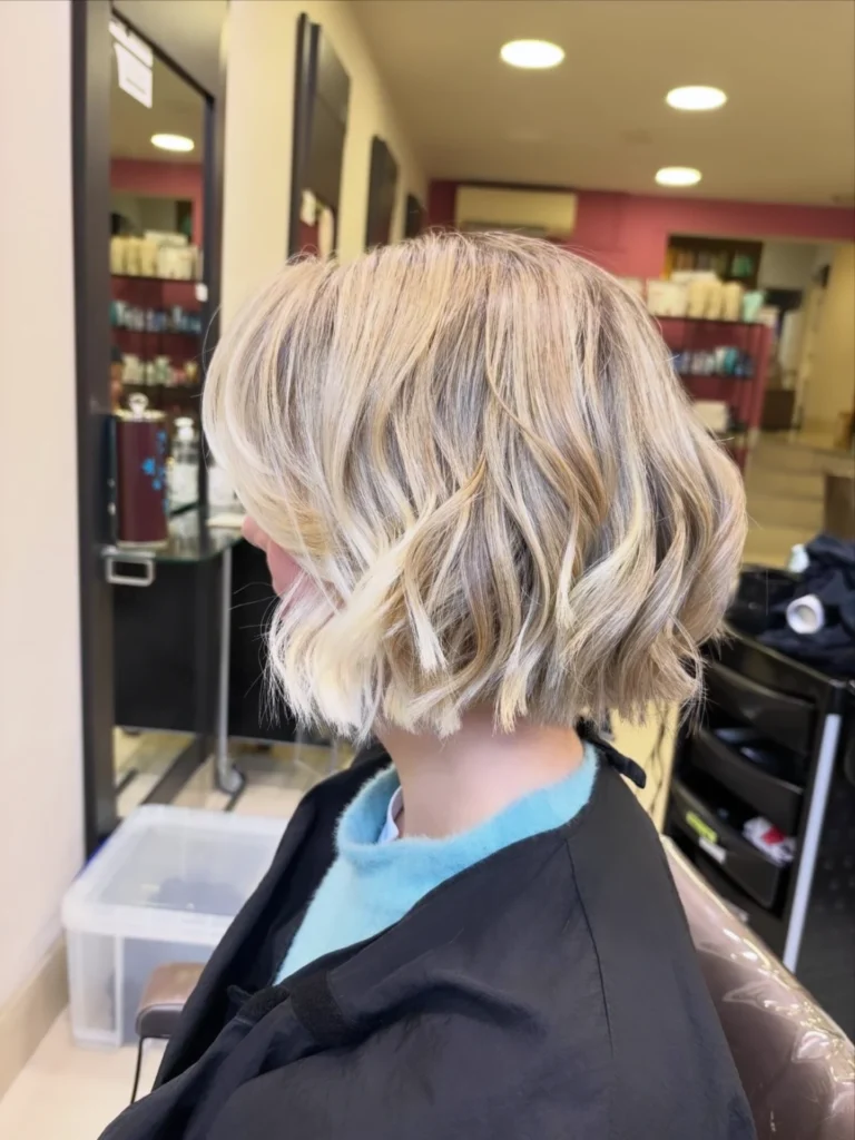 A woman in a salon chair viewed from the side profile showing off a heavily textured ash blonde bob with piecey waves.