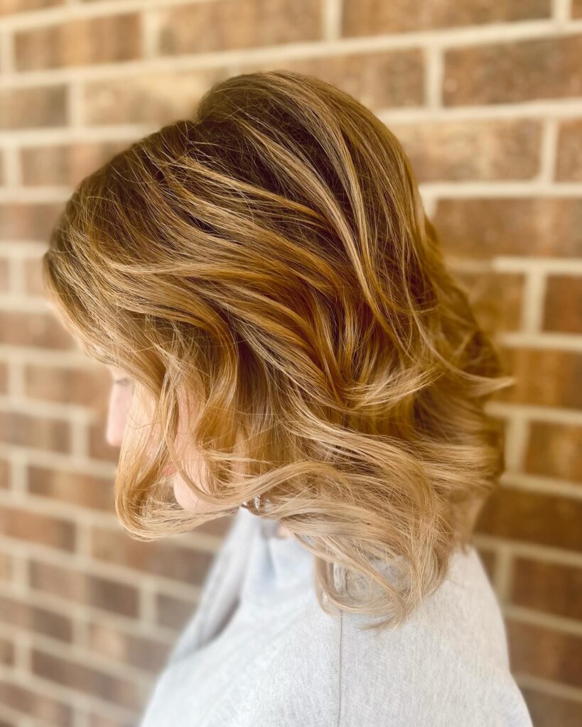 A woman shown in side profile against a brick wall sporting a golden honey blonde wavy long bob with voluminous choppy layers.