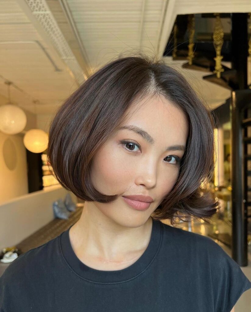 A woman looking directly at the camera showcasing a voluminous chin length brunette bob with a soft rounded flip styling.