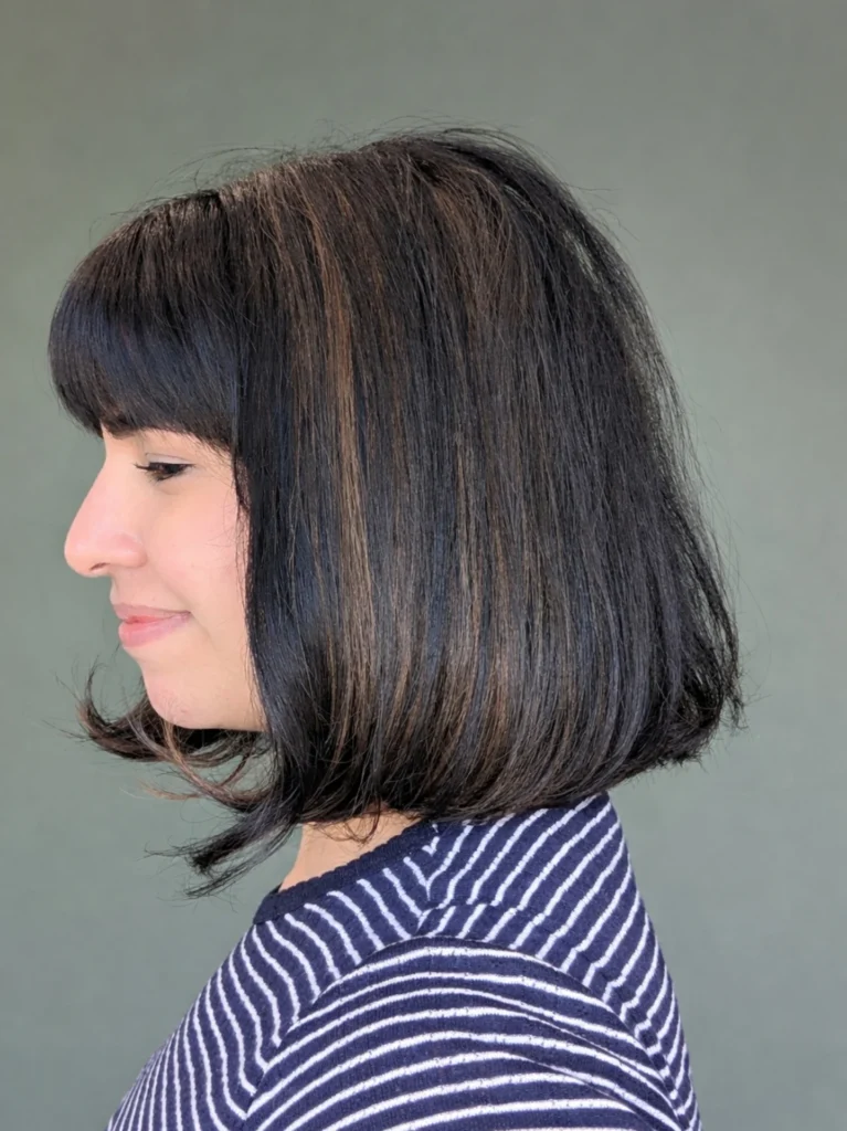 A woman showing the side profile of a chin length espresso bob with subtle bronde highlights and a full fringe.
