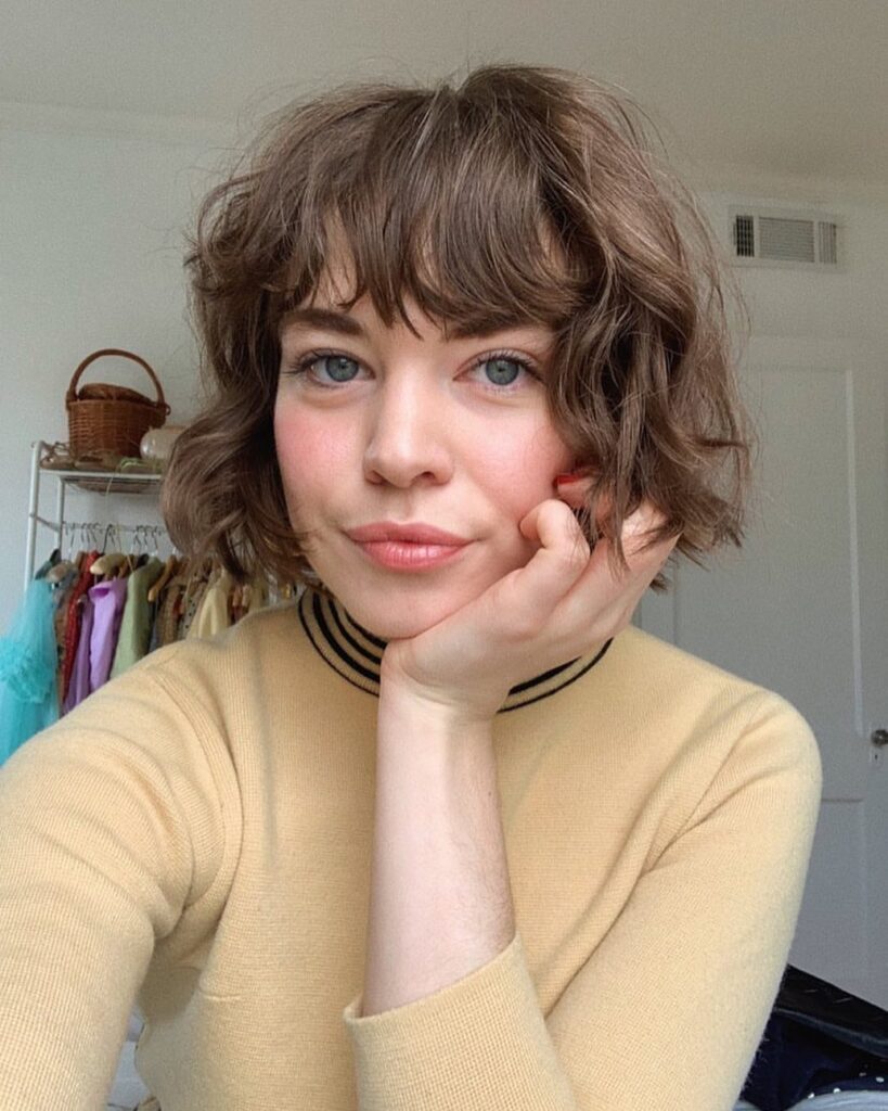 A woman with blue eyes resting her chin on her hand showcasing her wavy chin length light brown bob with choppy layers and full bangs.