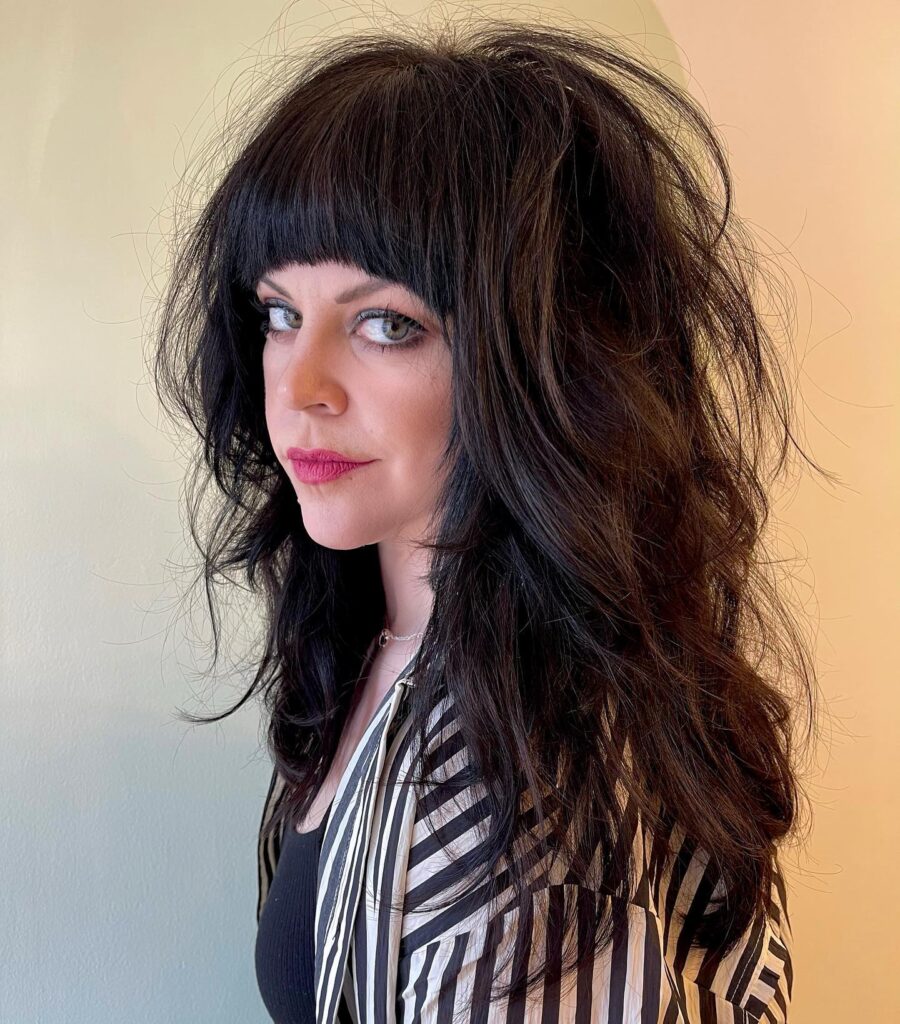 A woman with long, voluminous layered black shag hair featuring texturized waves and a thick blunt fringe.