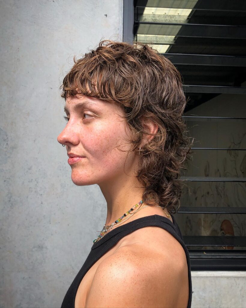 A person from a side profile view showcasing a curly, wet-look caramel brown mullet shag haircut with a short piecey fringe.