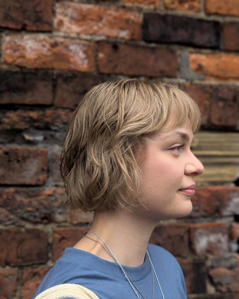 A side profile of a woman with a textured golden blonde choppy bob and soft side-swept bangs against a brick wall background.
