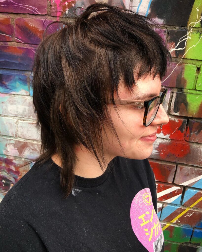 A person from a side profile view wearing glasses and a choppy, heavily layered brunette mixie shag haircut with short fringe.