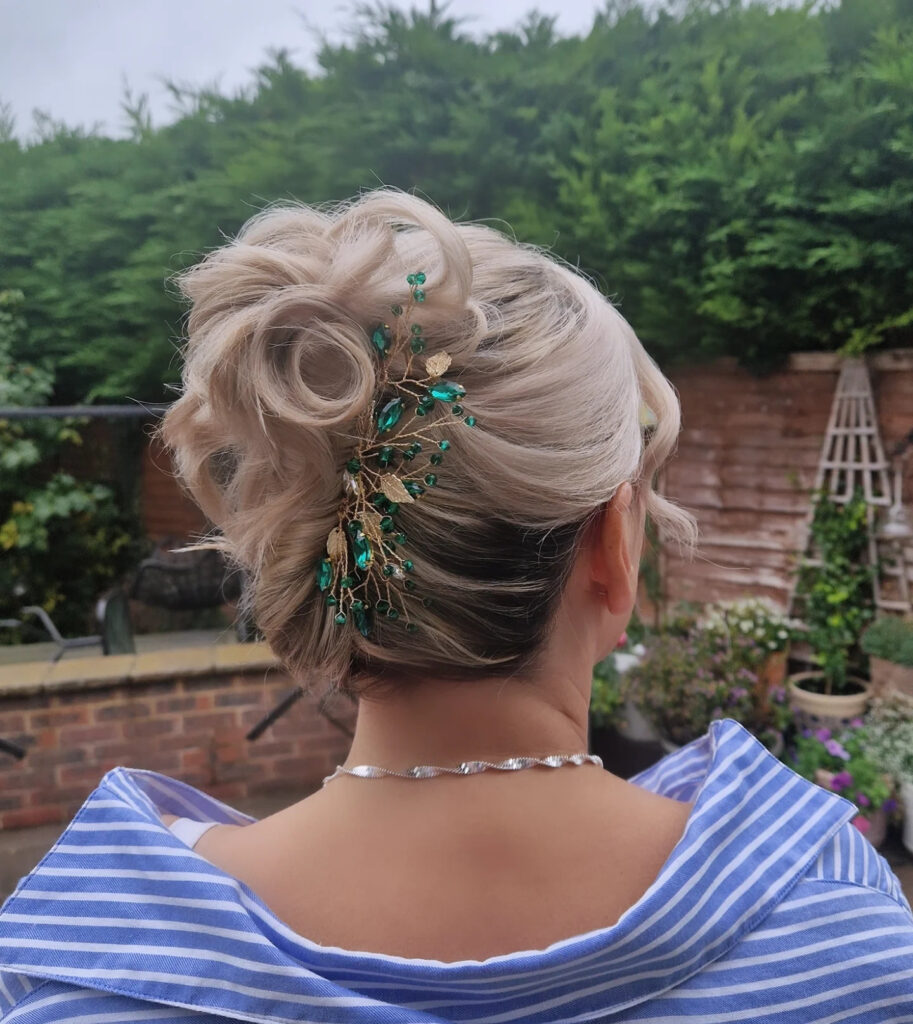 A back view of a woman with ash-blonde hair styled in a high, textured updo decorated with a gold-and-emerald-green jeweled hair vine.