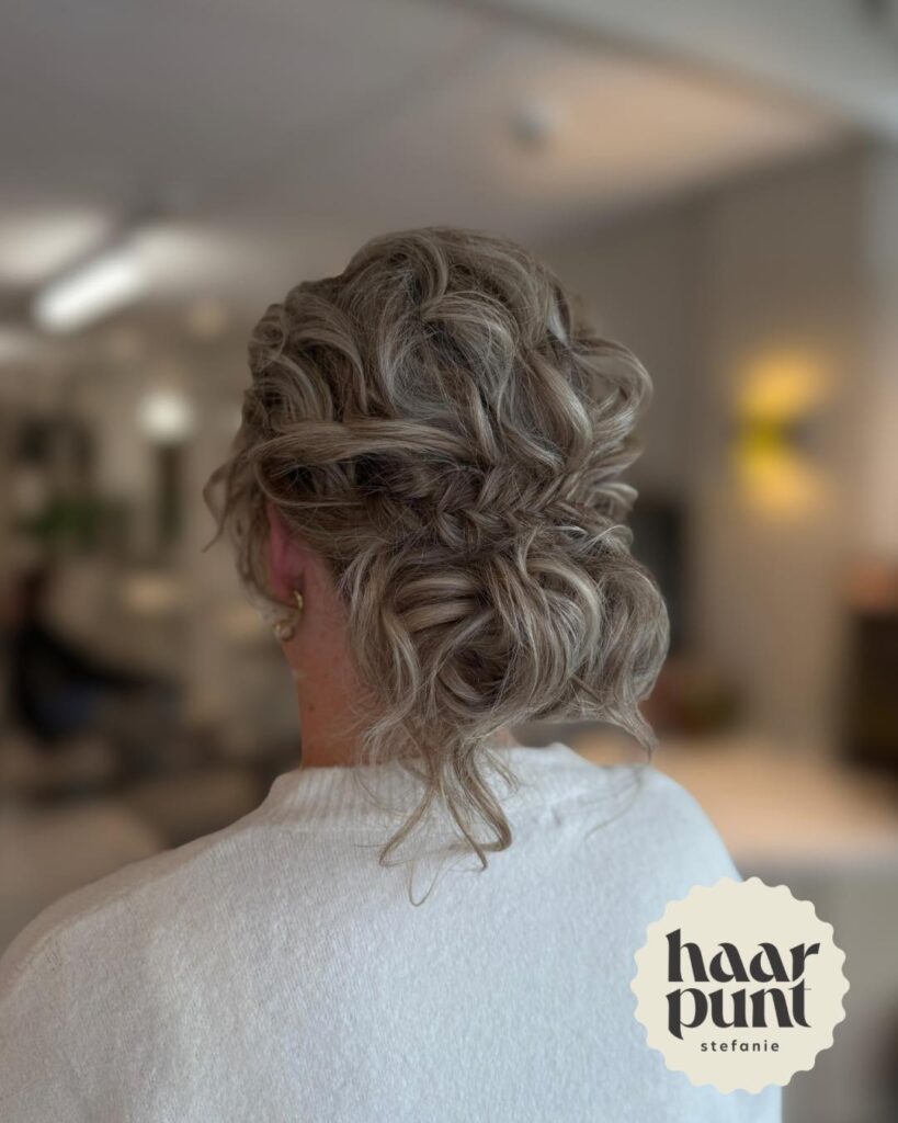 A back view of an ash-blonde curly bob styled into an updo with a fishtail braid detail and loose curls at the nape.