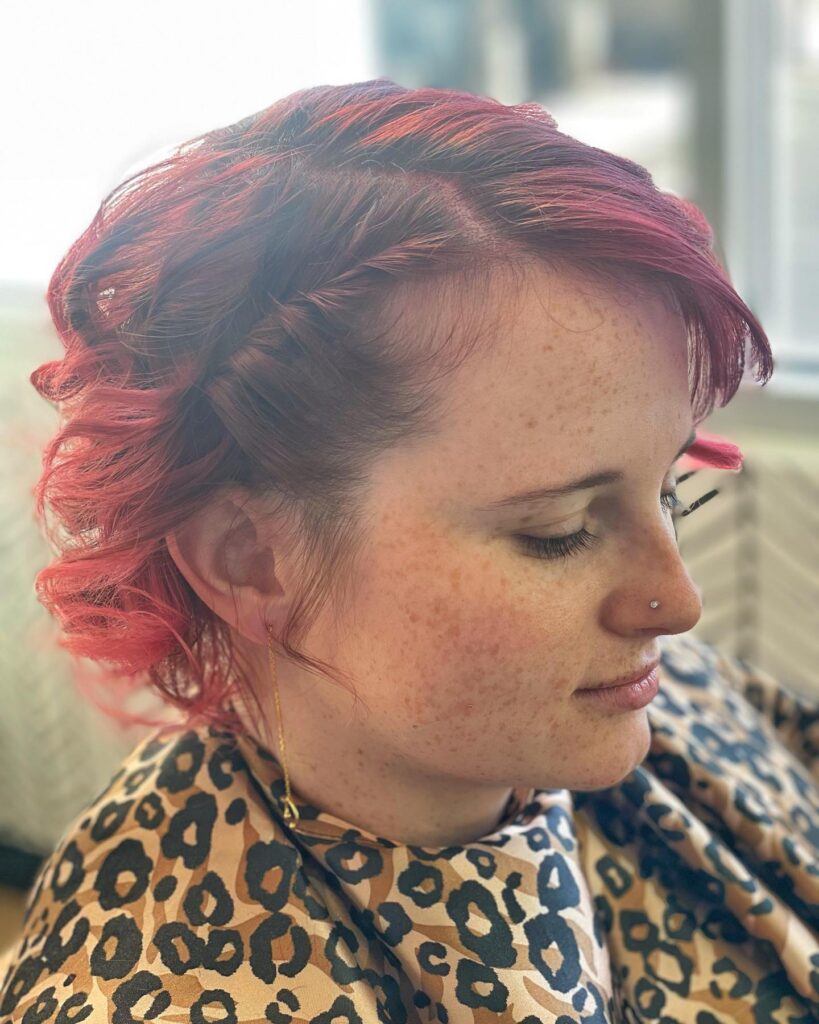 A woman with bright magenta curly hair styled with a side fishtail braid and voluminous ringlets.