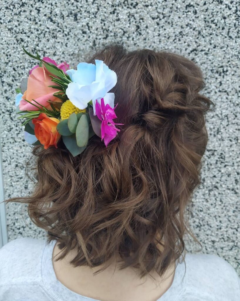 A back view of a woman with a wavy brunette bob styled in a half-updo accented with a large cluster of colorful tropical flowers.