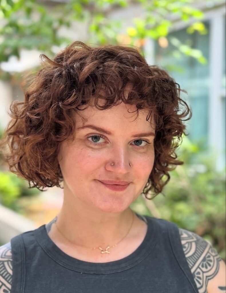 A woman with a curly auburn French bob and short curly bangs smiling at the camera.