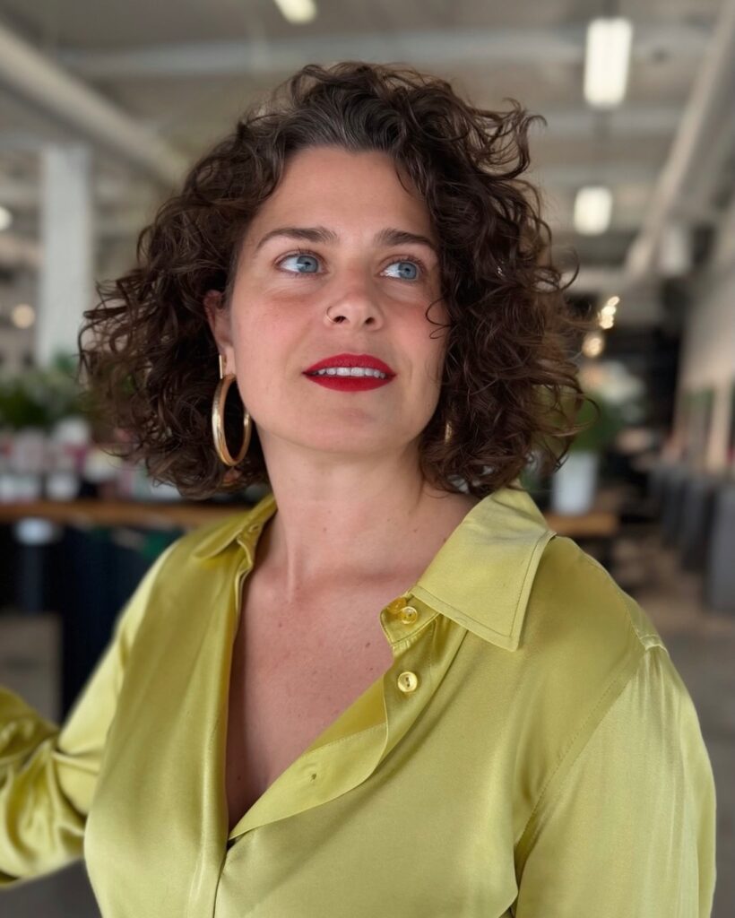 A woman with a side-parted, deep brown, curly French bob wearing gold hoop earrings and red lipstick.