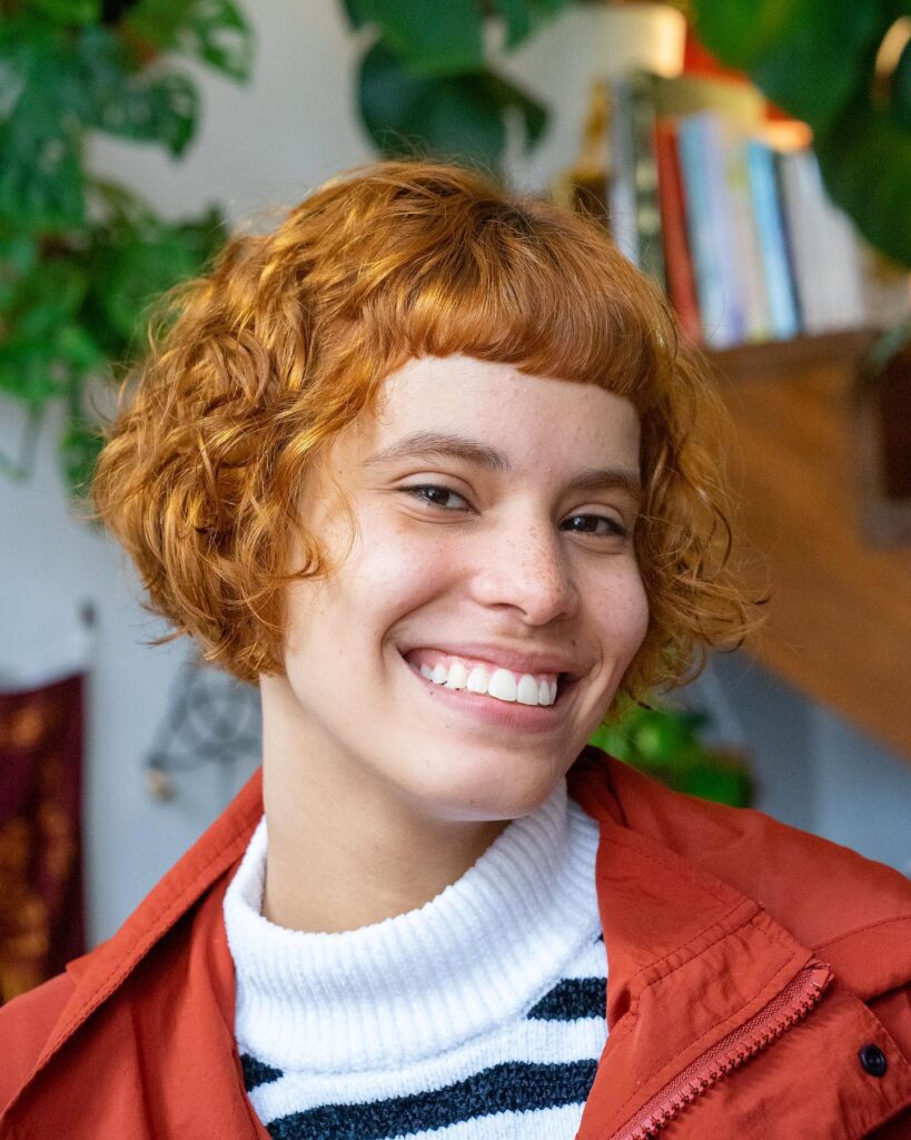 A smiling woman with a short, curly golden ginger French bob and blunt micro bangs.
