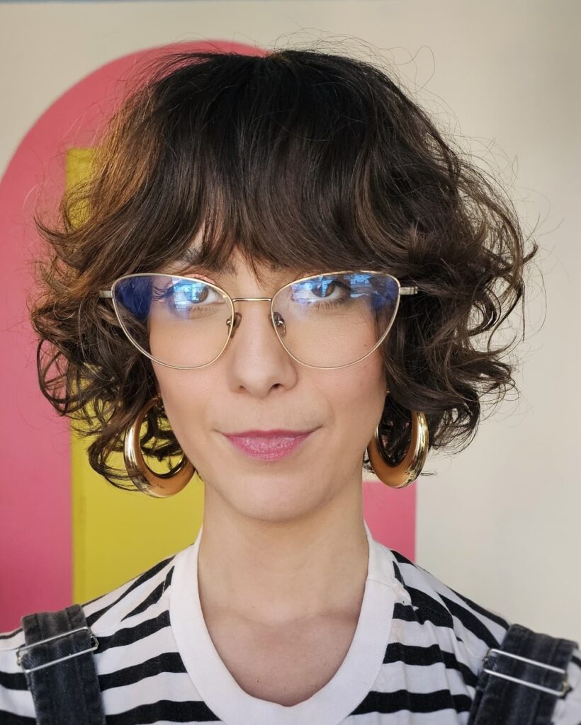 A woman with a dark brunette curly French bob featuring long wispy bangs and voluminous tousled layers.