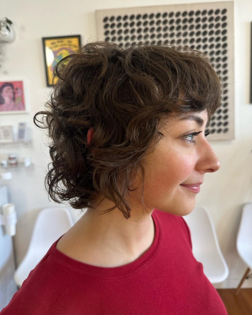 A side profile of a woman with a layered dark brown curly French bob and wispy bangs.
