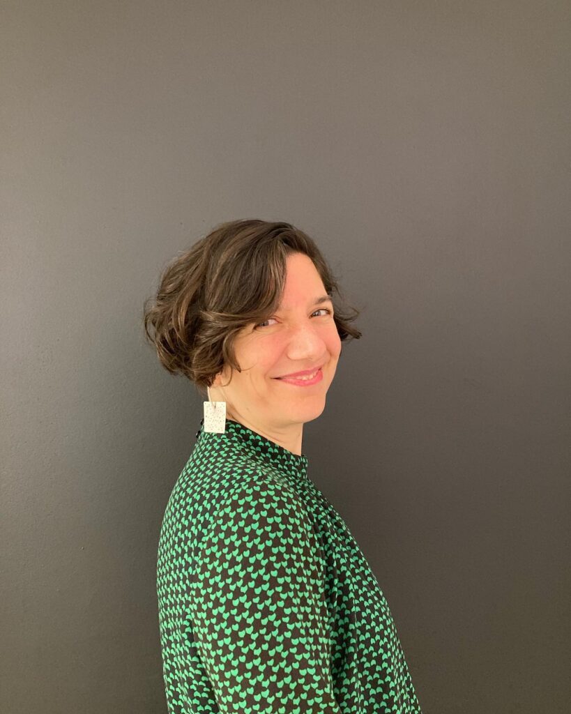 A woman with a short ash-brown wavy French bob and side-swept bangs smiling against a dark background.