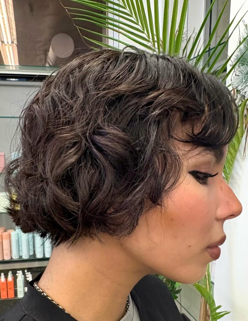 A profile view of a woman with a dark brunette curly French bob featuring short textured bangs and glossy waves.