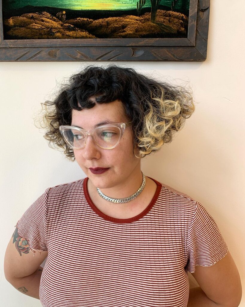 A woman with a two-tone black and blonde curly French bob and very short micro bangs.