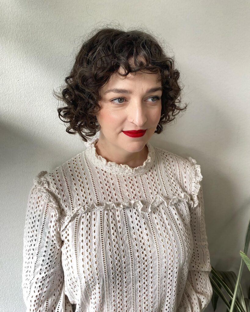 A woman with a cool brunette curly French bob featuring eyebrow-skimming bangs and bouncy jaw-length ringlets.