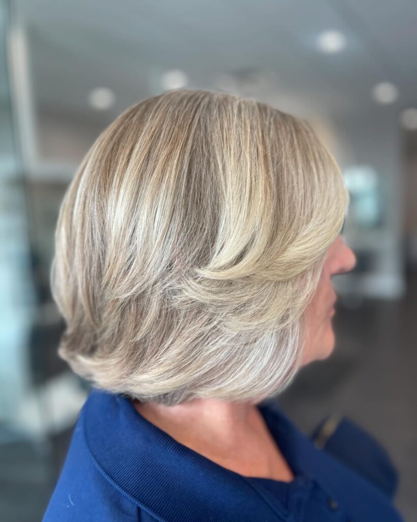 A woman with a neck-length champagne blonde layered bob featuring feathered side-swept tiers and a voluminous finish.