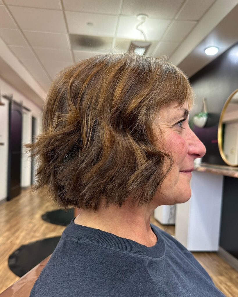 A woman in a salon showing a side profile of a jaw-length warm chestnut bob with caramel highlights and soft wavy texture.