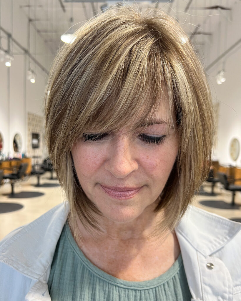A woman with a chin-length sandy blonde layered bob featuring a thick, wispy fringe and feathered side layers.