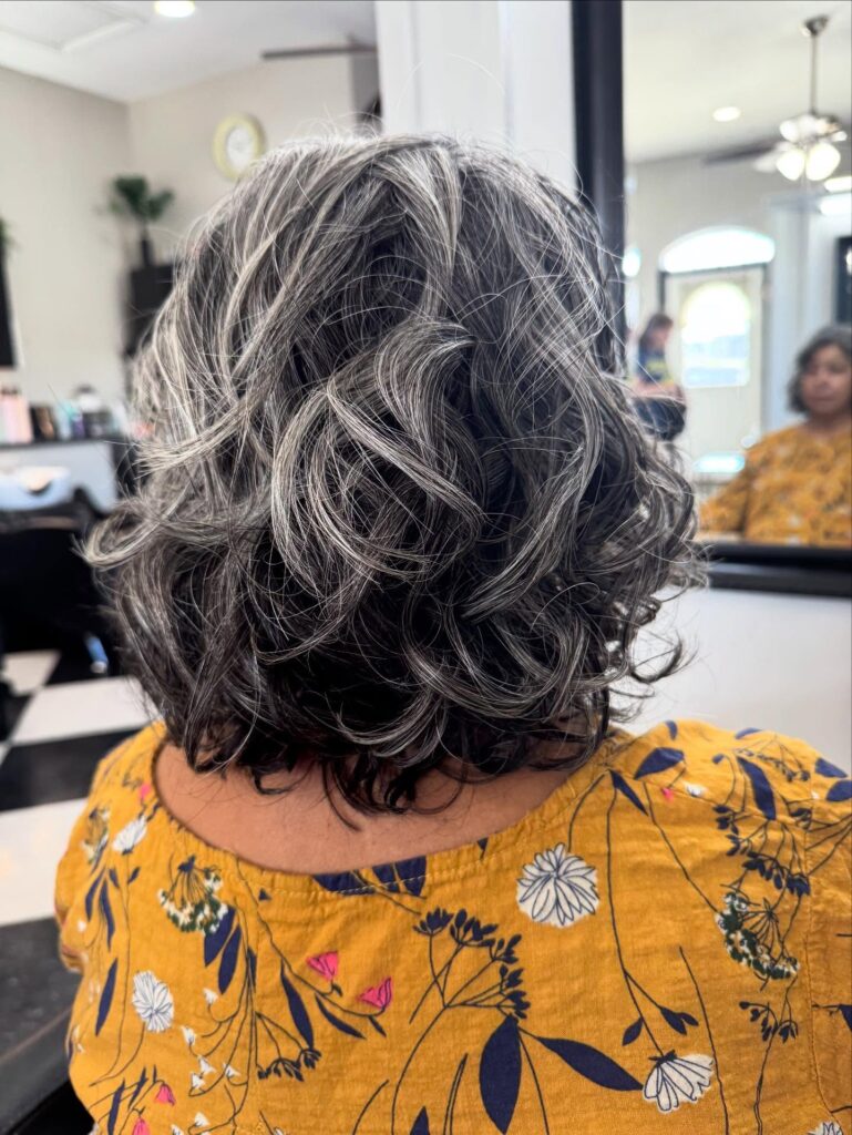 A woman from the back is showing a neck-length salt-and-pepper bob with defined curls and bright silver highlights.