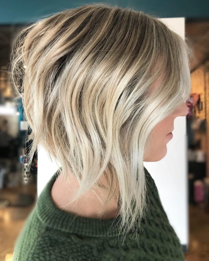 A side profile of a woman sporting a heavily angled long bob with wavy platinum blonde hair and dark roots.