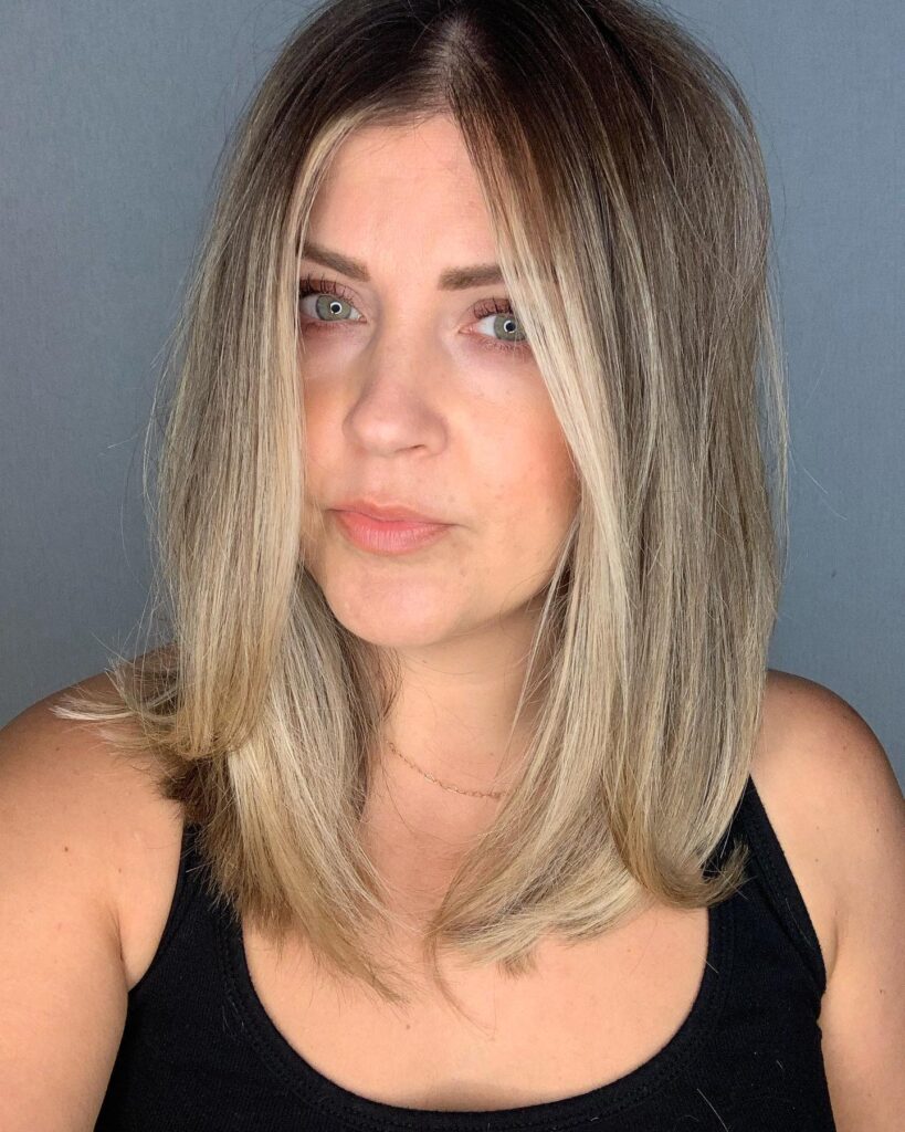 A woman with a middle-parted, angled, long bob featuring sandy blonde highlights and darker roots.