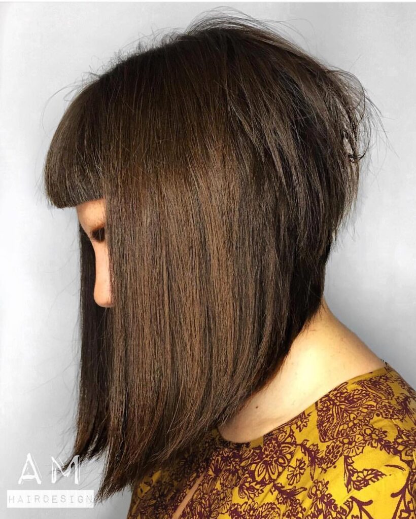 A side view of a woman sporting a steep-angled long brunette bob with a short micro fringe and voluminous layers at the back.