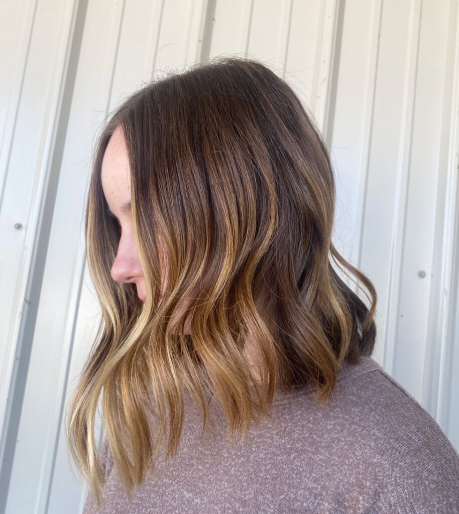 A woman with a chestnut brown angled long bob featuring warm caramel balayage highlights and soft wavy texture.