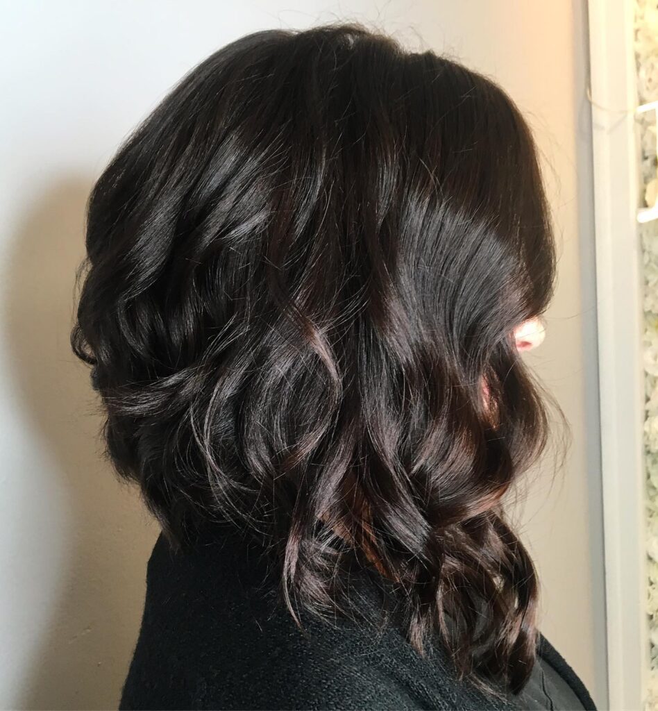 A woman with deep mahogany brown hair styled in a steeply angled long bob with thick, bouncy curls.