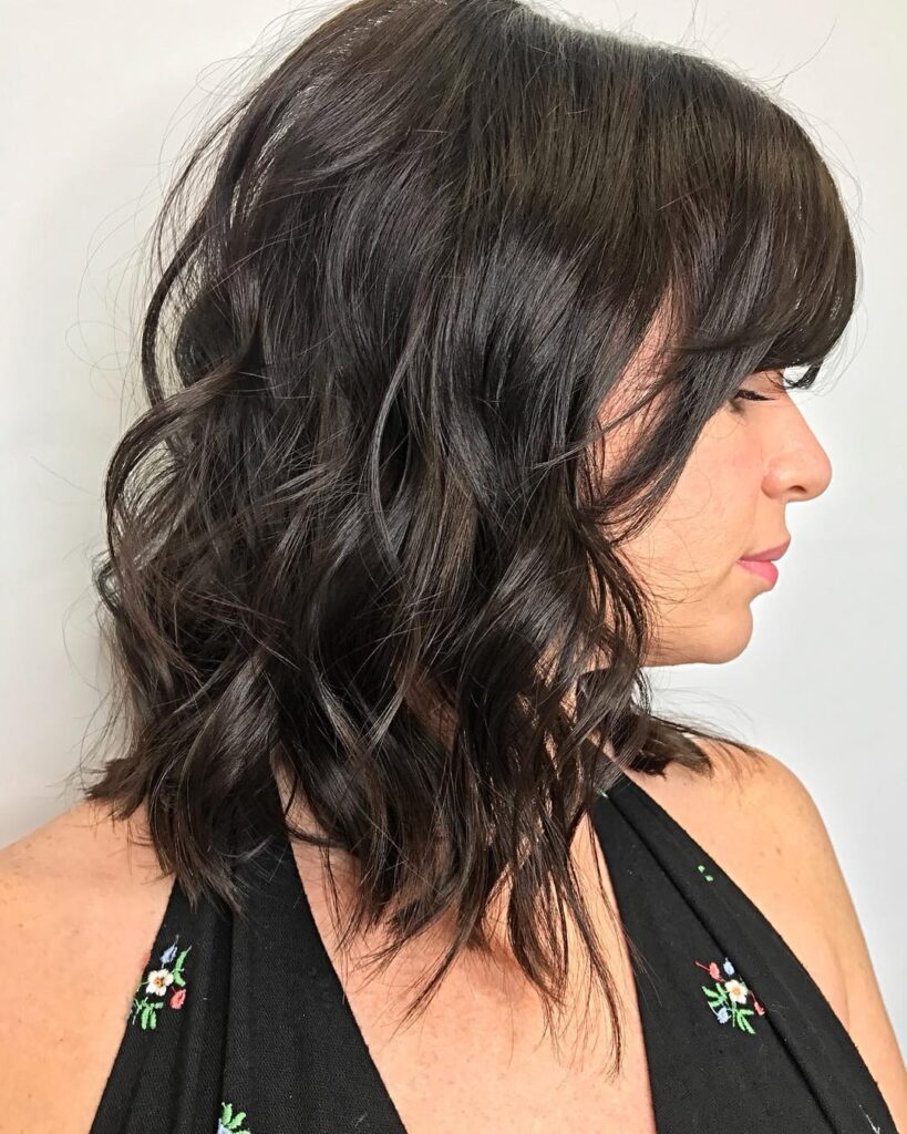 A side profile of a woman with a wavy dark brunette angled lob featuring choppy layers and side-swept bangs.