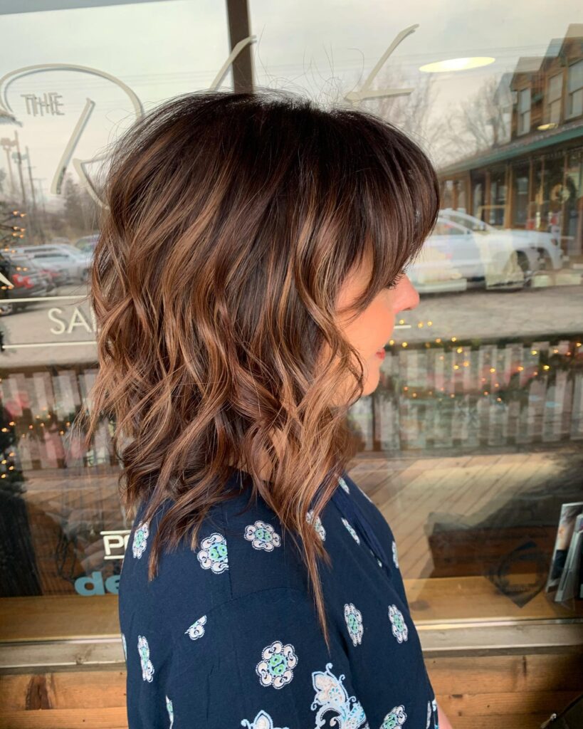 A side profile of a woman with an angled long bob featuring copper balayage, shaggy layers, and a full wispy fringe.