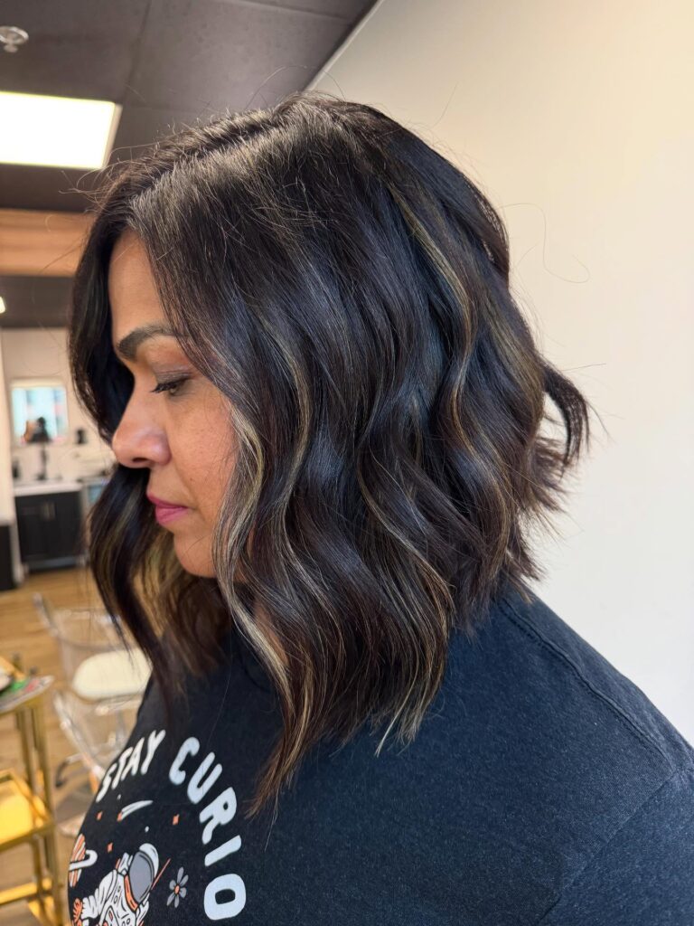 A woman with dark brown wavy hair in an angled lob featuring thin blonde highlights and choppy ends.
