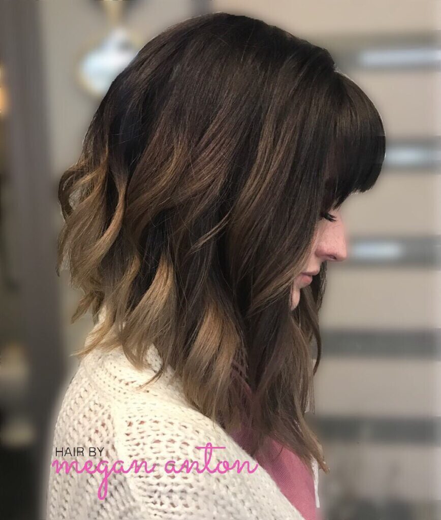 A side profile of a woman with an angled long bob featuring a brown-to-blonde ombre and a full straight fringe.