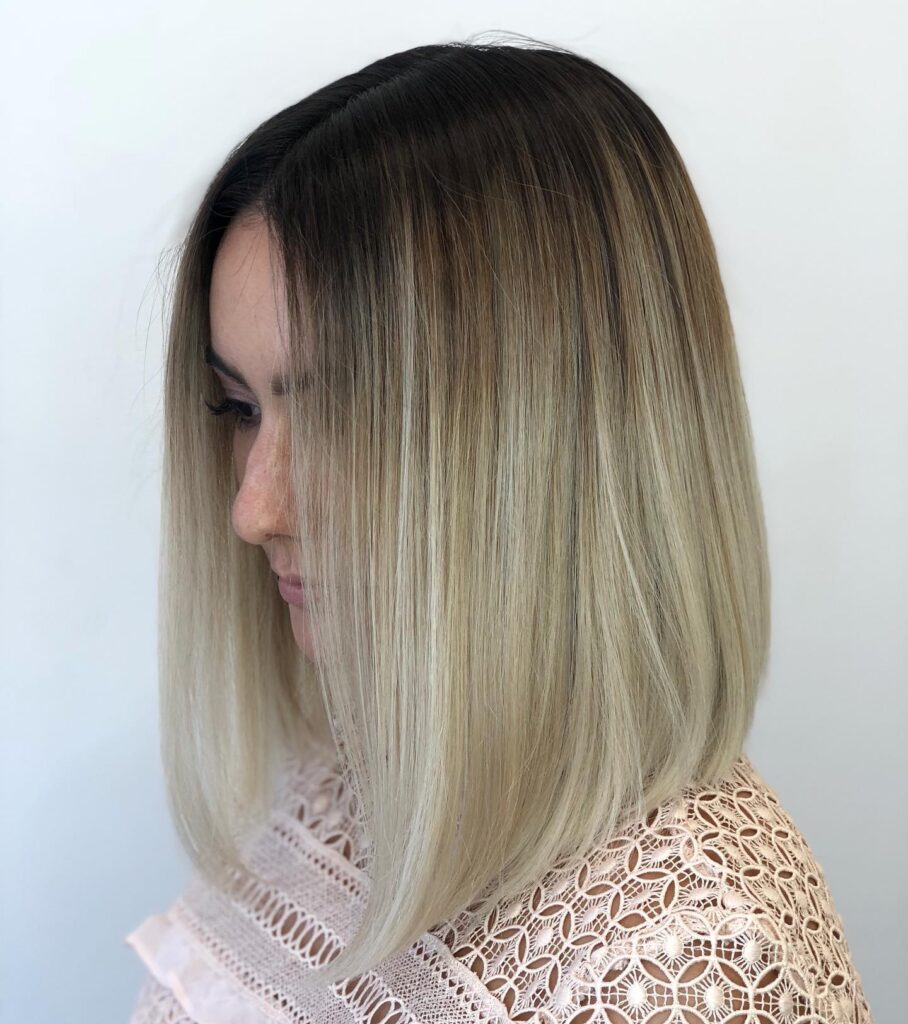 A side profile of a woman with a straight-angled long bob featuring dark roots and creamy blonde ends.