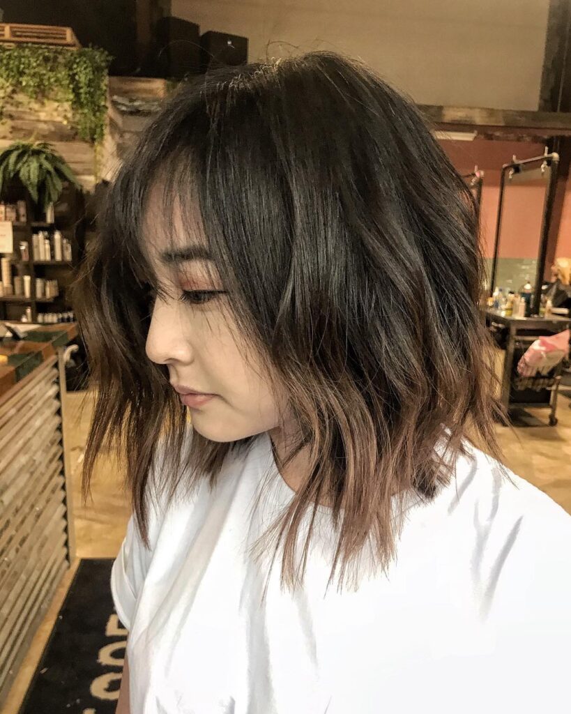 A side profile of a woman with a wavy mocha-angled long bob featuring wispy bangs and caramel-toned ends.