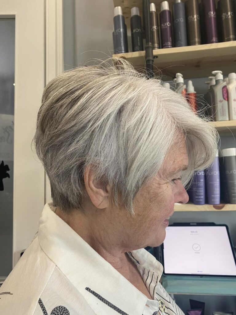 A woman in profile wearing a feathered ashy-grey pixie cut with soft layers and side-swept bangs.