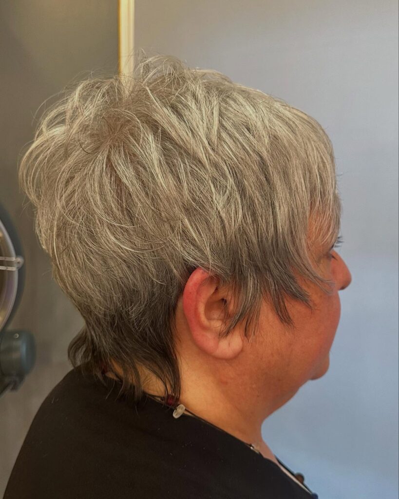 A side view of a woman with a salt-and-pepper pixie cut featuring shaggy layers and a wispy neckline.