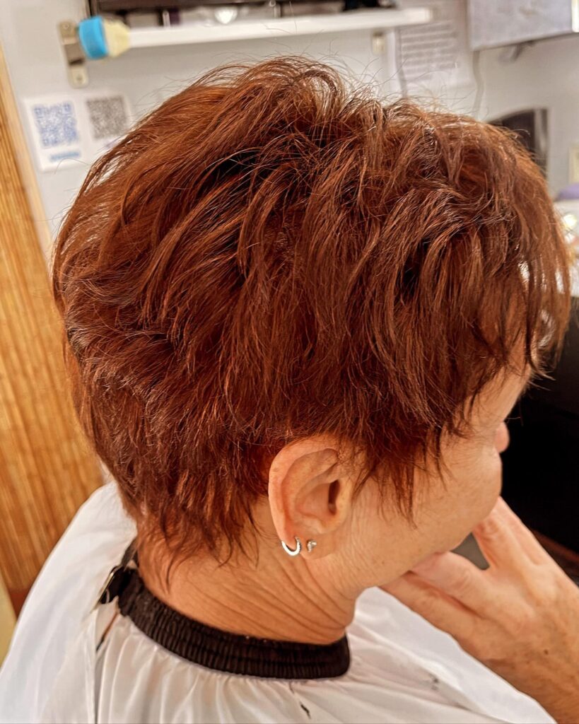A woman over 60 with a short texturized auburn pixie cut featuring wispy layers and a tapered neckline.