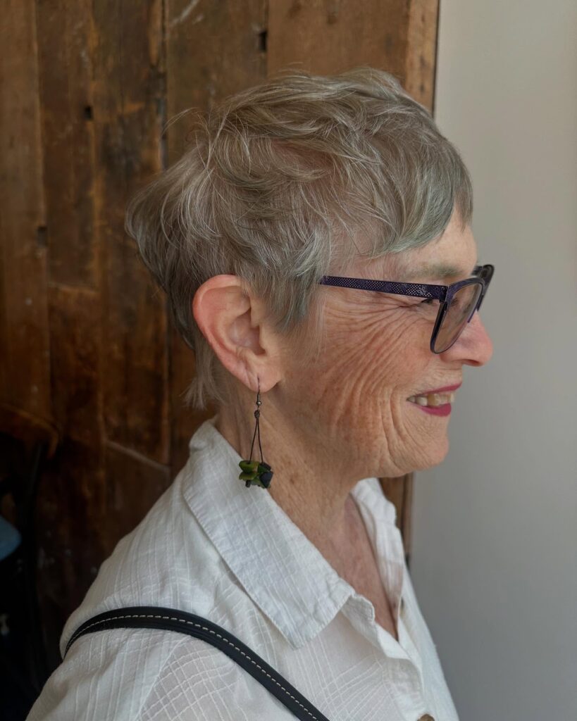 A woman over 60 with a short wispy silver pixie cut featuring micro bangs and soft layers around the ears.