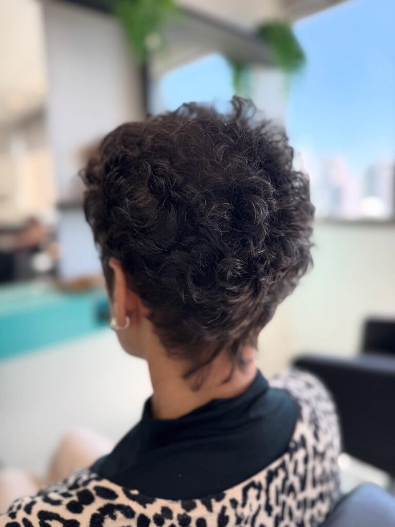 A woman over 60 is seen from the back with a short curly pixie cut in a charcoal salt-and-pepper shade featuring a tapered neckline.