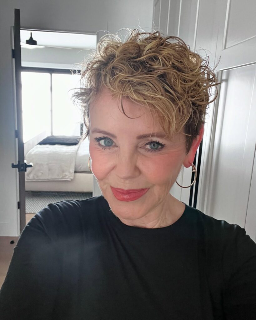 A woman smiling with a golden blonde curly pixie cut that features voluminous layers on top.