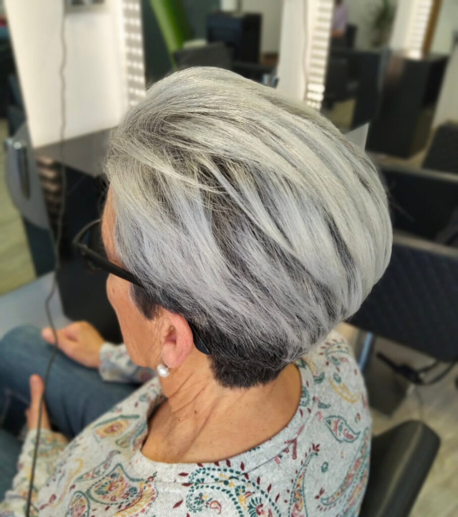 A woman from a side-back angle with a voluminous silver pixie cut featuring dark shadow roots and swept-back layers.